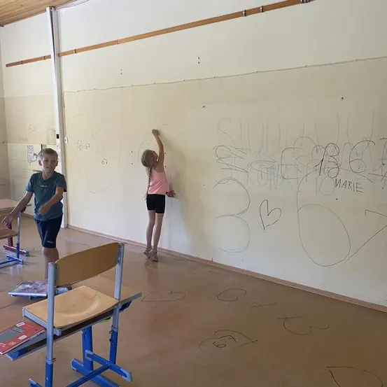Zwei Kinder zeichnen auf eine große weiße Wand in einem Klassenzimmer. Die Wand hat verschiedene Zeichnungen und Schriften darauf. Ein Junge steht in der Nähe eines Schreibtisches, und ein Mädchen steht auf einem Stuhl.