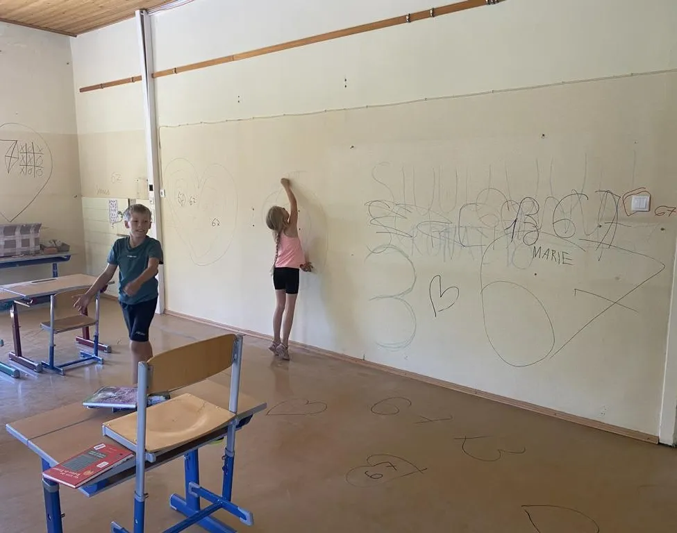 Zwei Kinder zeichnen auf eine große weiße Wand in einem Klassenzimmer. Die Wand hat verschiedene Zeichnungen und Schriften darauf. Ein Junge steht in der Nähe eines Schreibtisches, und ein Mädchen steht auf einem Stuhl.