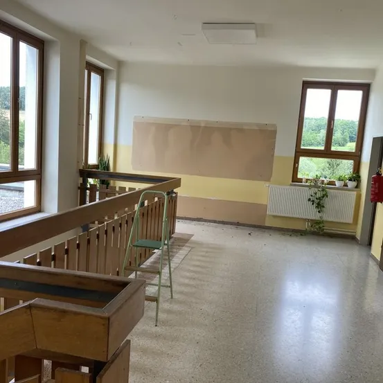 Ein leerer Raum mit einem hölzernen Geländer, einem grünen Stuhl und einem Feuerlöscher. Der Raum hat weiße Wände, eine weiße Decke und eine gelbe Wand mit einem Heizkörper und Pflanzen.