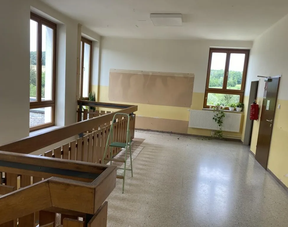 Ein leerer Raum mit einem hölzernen Geländer, einem grünen Stuhl und einem Feuerlöscher. Der Raum hat weiße Wände, eine weiße Decke und eine gelbe Wand mit einem Heizkörper und Pflanzen.