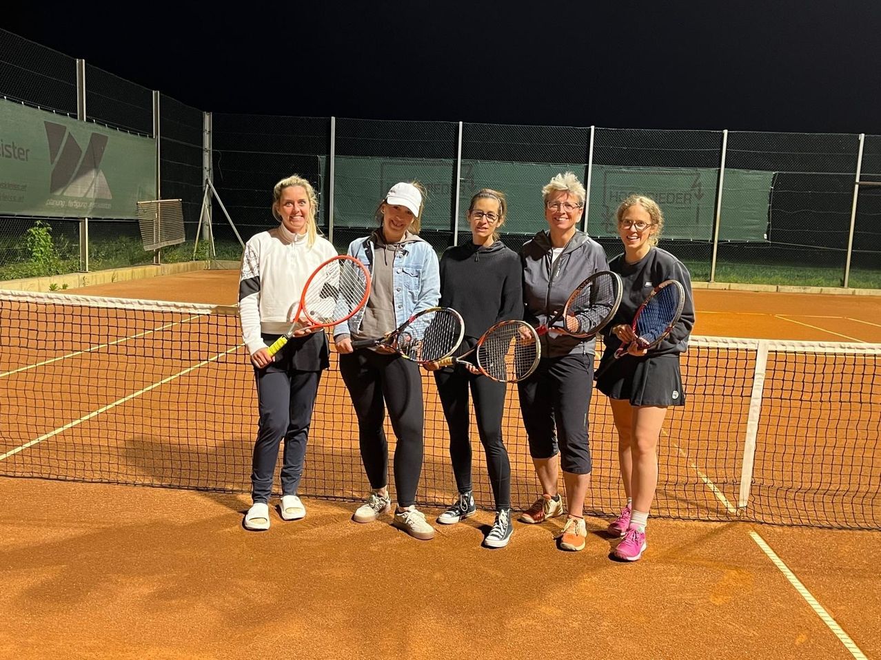 Fünf Frauen stehen auf einem Tennisplatz, lächeln und halten Tennisschläger in den Händen. Sie tragen Sportbekleidung und Schuhe. Hinter ihnen ist ein Netz und ein Zaun. Der Platz ist von Licht beleuchtet.