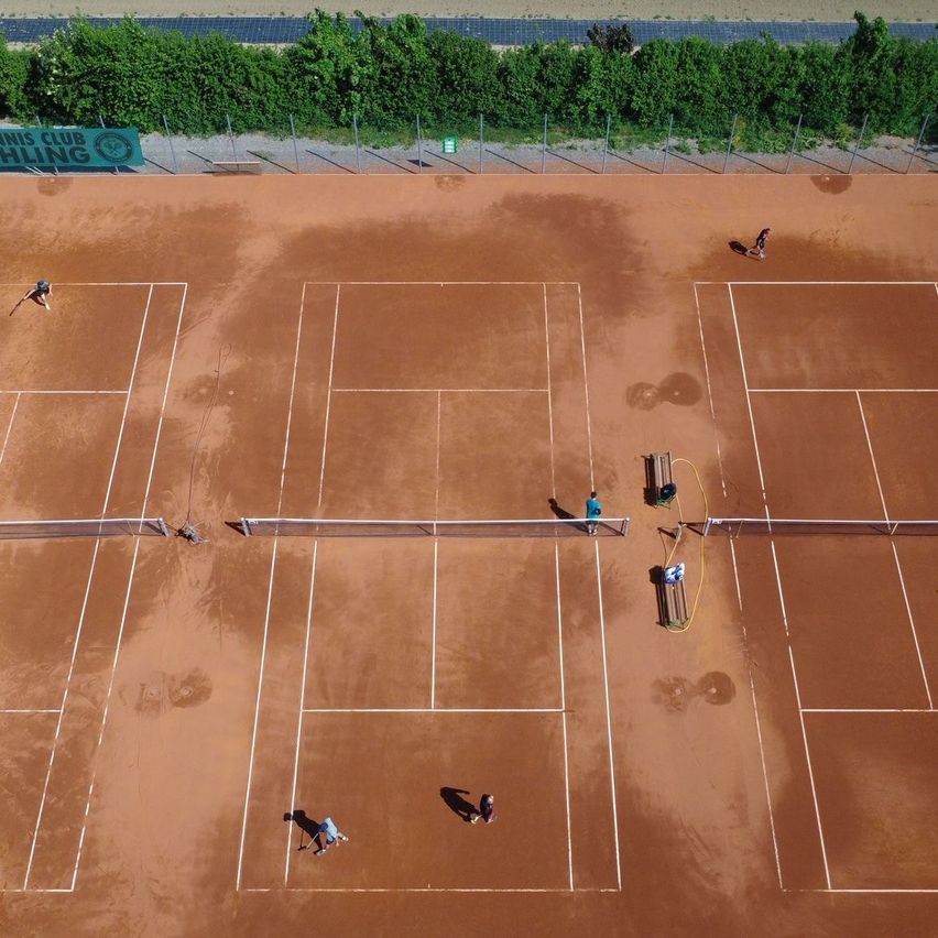Luftaufnahme eines Tennisplatzes mit spielenden Personen, einem grünen Schild und Bänken.