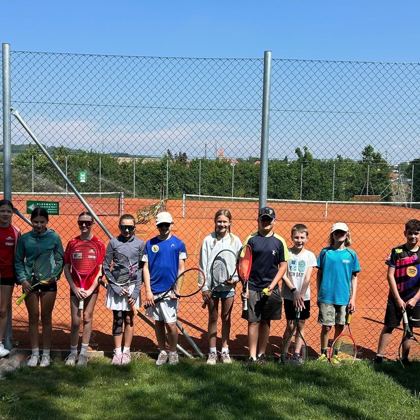 Eine Gruppe junger Leute mit Tennisschlägern steht vor einem Tennisplatz und posiert für ein Foto. Sie alle lächeln. Dahinter trennt ein Maschendrahtzaun den Platz vom restlichen Bereich.