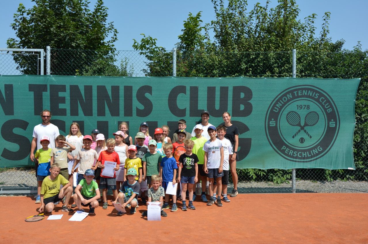 Eine Gruppe von Kindern und Erwachsenen posiert für ein Foto auf einem Tennisplatz, hält Urkunden und trägt Mützen. Der Hintergrund zeigt ein grünes Banner mit den Worten Tennis Club und einem Logo.