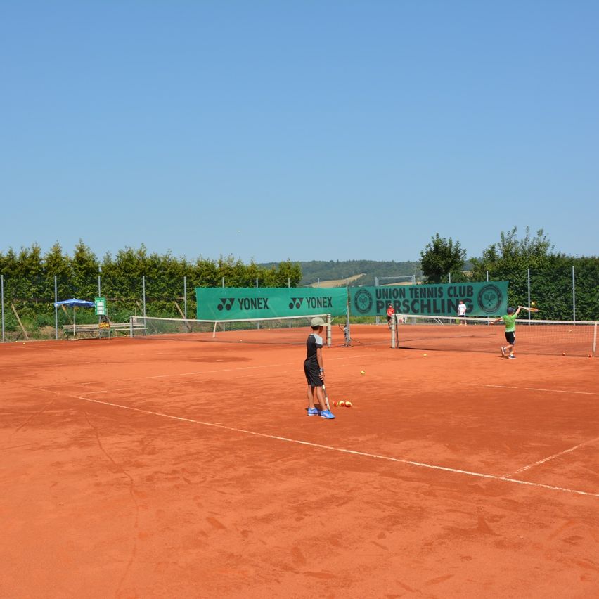 Ein Tennisplatz mit Spielern im Training, einer hält Schläger und Ball, andere spielen auf gegenüberliegenden Seiten, mit einem grünen Banner im Hintergrund, das 'UNION TENNIS CLUB PERSCHLINGS' steht.