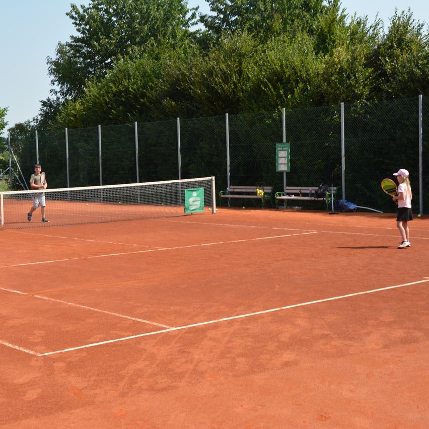 Zwei Personen spielen Tennis auf einem Sandplatz mit Netz, umgeben von einem Zaun und Bäumen.