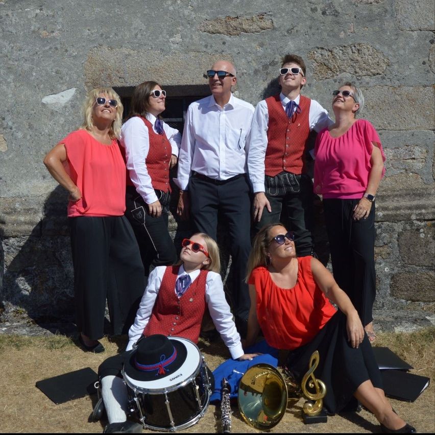 Eine Gruppe von Menschen, die in passenden Westen und Sonnenbrillen gekleidet sind, posiert für ein Foto vor einer Steinmauer. Musikinstrumente liegen auf dem Boden verstreut.
