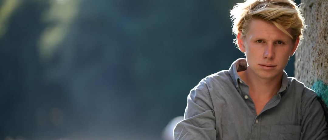 Ein Mann mit blondem Haar steht mit verschränkten Armen in einem grauen Hemd. Er hat Armbänder an seinem Handgelenk und schaut nach rechts. Der Hintergrund ist verschwommen.