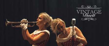 Zwei Frauen spielen auf der Bühne in roten gepunkteten Kleidern, eine spielt Trompete und die andere singt in ein Mikrofon. Im Hintergrund ist eine Gitarre zu sehen. Der Text 'VINT Duet' befindet sich oben im Bild.