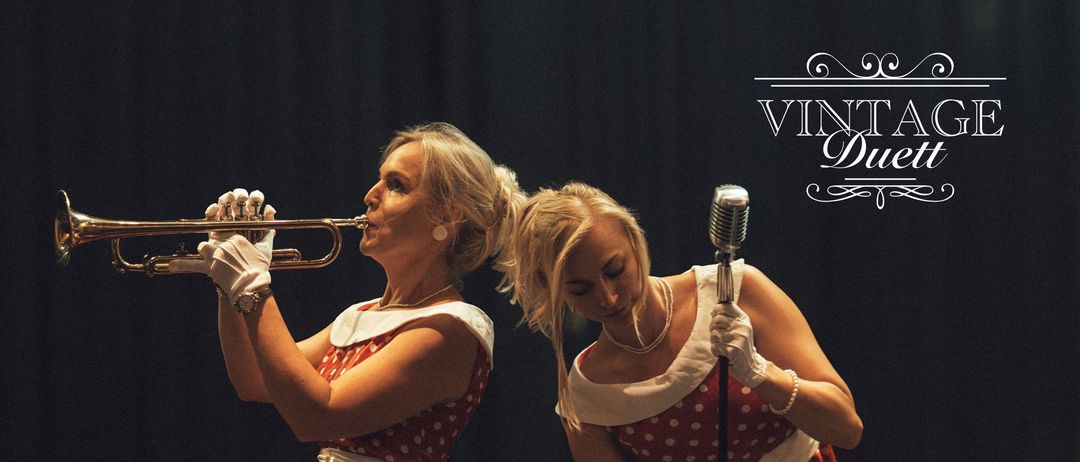 Zwei Frauen spielen auf der Bühne in roten gepunkteten Kleidern, eine spielt Trompete und die andere singt in ein Mikrofon. Im Hintergrund ist eine Gitarre zu sehen. Der Text 'VINT Duet' befindet sich oben im Bild.