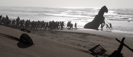 Eine Gruppe von Menschen zieht eine große pferdeähnliche Statue zum Meer auf einem Strand. Die Statue steht auf einer Plattform mit Rädern und wird von Seilen gezogen.