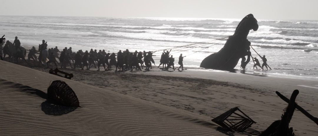 Eine Gruppe von Menschen zieht eine große pferdeähnliche Statue zum Meer auf einem Strand. Die Statue steht auf einer Plattform mit Rädern und wird von Seilen gezogen.