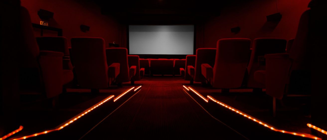 Ein leeres Kino mit roten Stühlen und einer großen leeren Leinwand. Rote Lichter beleuchten den Gang.