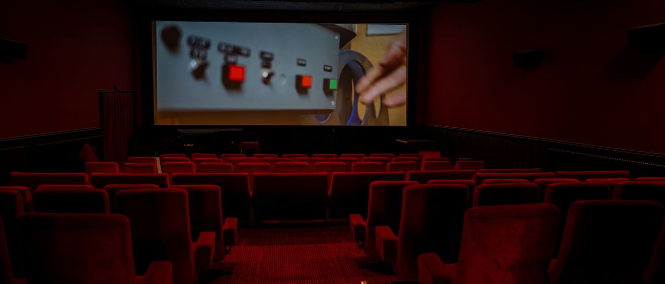 Ein leeres Kino mit roten Stühlen und einer großen Projektionsleinwand, die einen Film zeigt. Die Leinwand zeigt eine Hand, die einen Filmrollen einstellt.