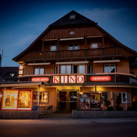 Bei Nacht ist das Äußere eines Filmcasino Kino Groebming beleuchtet. Es hat einen Balkon, Filmplakate und Tische draußen.