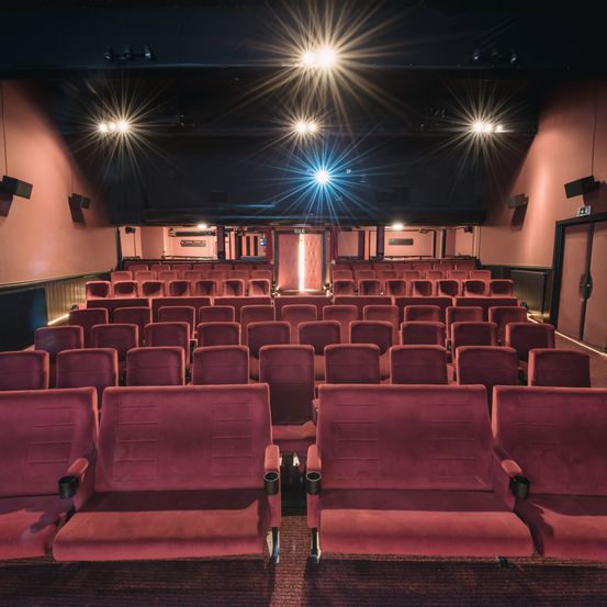 Ein leeres Kino mit roten Sitzen, beleuchtet von mehreren Scheinwerfern an der Decke, und zwei offenen Türen am Ende.