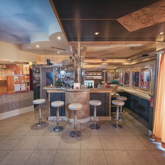 Ein Barbereich mit Theke, Hockern, Regalen und Flaschen. An der Wand befinden sich auch Bilderrahmen.