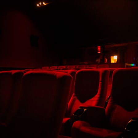 Ein schwach beleuchtetes Kino mit roten Sitzen, die in Reihen angeordnet sind, beleuchtet von einem einzelnen roten Licht. Der Raum ist dunkel, nur wenige Lichter an der Decke sind an.