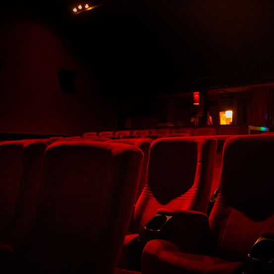 Ein schwach beleuchtetes Kino mit roten Sitzen, die in Reihen angeordnet sind, beleuchtet von einem einzelnen roten Licht. Der Raum ist dunkel, nur wenige Lichter an der Decke sind an.