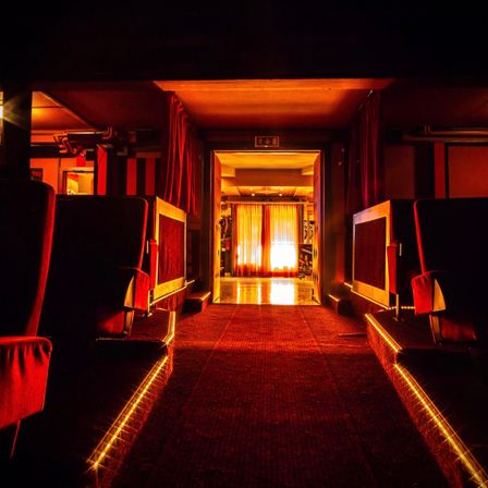 Ein schwach beleuchteter Korridor mit roten Stühlen auf beiden Seiten, roten Vorhängen und einem hellen gelben Licht am Ende.