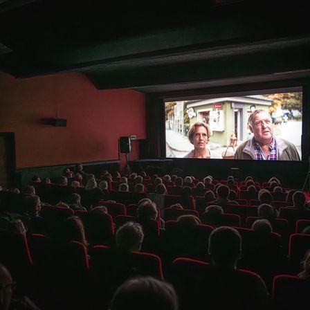 Ein Kino zeigt einen Film auf einer großen Leinwand mit zwei Personen, während das Publikum in Reihen roter Stühle zusieht.