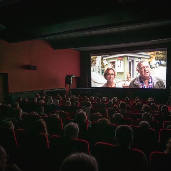 Ein Kino zeigt einen Film auf einer großen Leinwand mit zwei Personen, während das Publikum in Reihen roter Stühle zusieht.