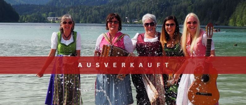 Fünf Frauen stehen lächelnd im Wasser und halten Musikinstrumente in der Hand. Sie tragen traditionelle Kleidung. Das Wort 'AUSVERKAUFT' ist rot auf dem Bild geschrieben. Hinter ihnen befindet sich ein See mit Blick auf die Berge.