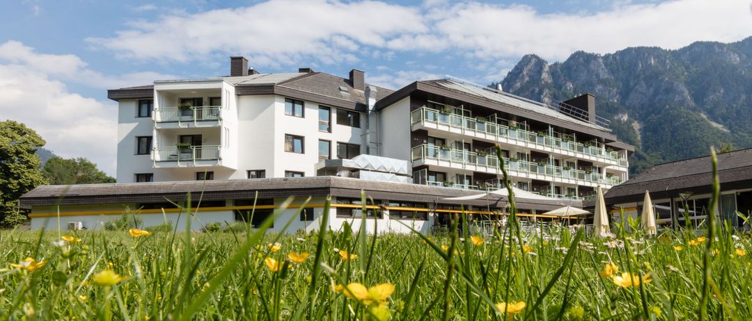 Ein modernes Hotel mit mehreren Stockwerken und Balkonen befindet sich auf einem Grasfeld mit gelben Blumen unter einem blauen Himmel mit Wolken und Bergen in der Ferne.