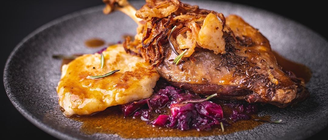 Ein Gourmet-Teller mit einem gebratenen Entenschenkel, bedeckt mit knusprigen Zwiebelringen und Kräutern, serviert mit Kartoffelpüree und Rotkohl.