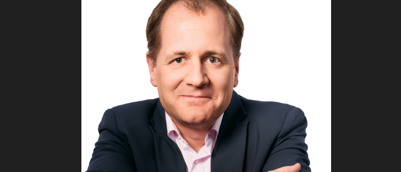 Ein Mann in einem dunklen Anzug und einem rosa Hemd, lächelnd, mit verschränkten Armen, vor einem weißen Hintergrund.