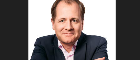 Ein Mann in einem dunklen Anzug und einem rosa Hemd, lächelnd, mit verschränkten Armen, vor einem weißen Hintergrund.