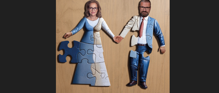 Ein Paar steht auf einem hölzernen Hintergrund, gekleidet in Puzzleteil-Kostüme, und hält Händchen. Der Mann trägt einen blauen Anzug und eine Krawatte, während die Frau ein blaues Kleid trägt.