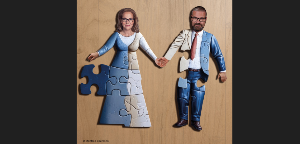 Ein Paar steht auf einem hölzernen Hintergrund, gekleidet in Puzzleteil-Kostüme, und hält Händchen. Der Mann trägt einen blauen Anzug und eine Krawatte, während die Frau ein blaues Kleid trägt.