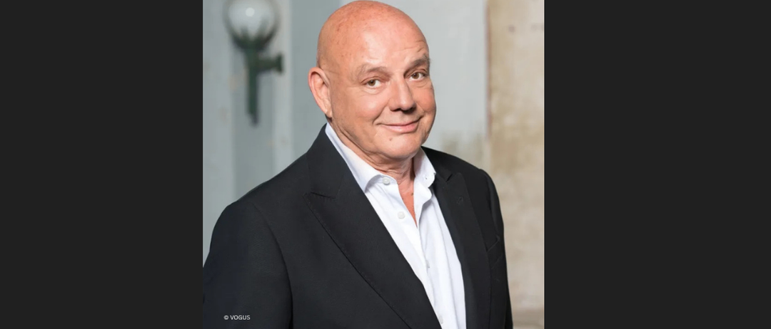 Ein Mann steht lächelnd und trägt ein weißes Hemd und eine schwarze Jacke. Er befindet sich in einem Studio mit unscharfem Hintergrund. Im Hintergrund befindet sich eine Lampe.