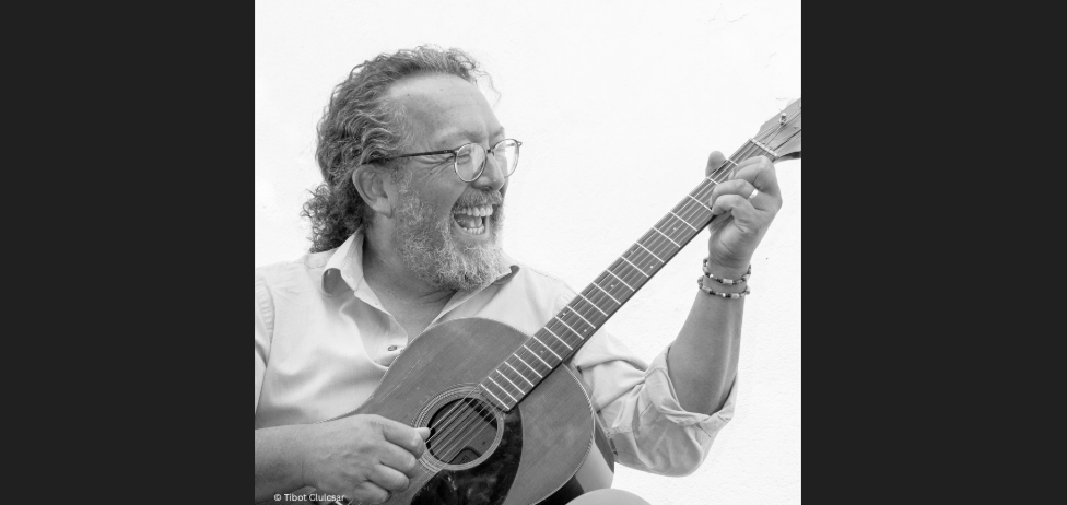 Ein schwarz-weißes Porträt eines bärtigen Mannes, der eine Gitarre spielt. Er trägt eine Brille und ein Armband.