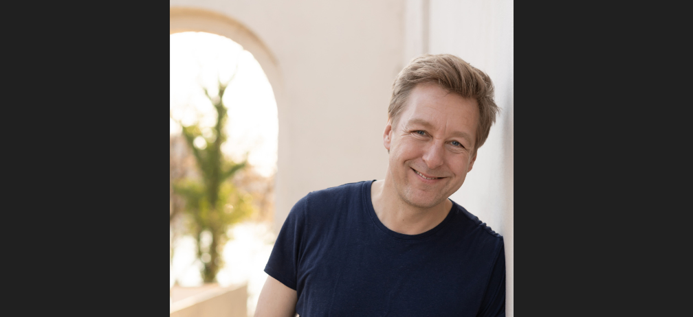Ein Mann mit einem Lächeln lehnt sich gegen eine weiße Wand und trägt ein blaues T-Shirt. Dahinter ist ein verschwommenes Bild eines Baumes und eines Bogens zu sehen. Das Foto wurde von Helge Bauer aufgenommen.