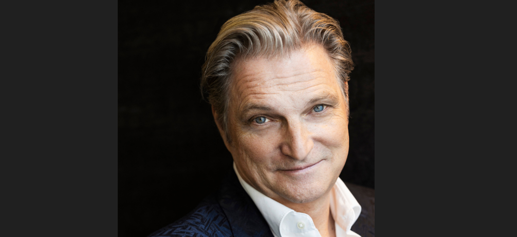 Ein Porträt eines älteren Mannes mit grauem Haar und blauen Augen, der ein weißes Hemd und eine blaue Jacke trägt und lächelt. Er hat Falten auf seiner Stirn und um seine Augen. Er schaut nach vorne. Der Hintergrund ist dunkel. Das Wasserzeichen ist unten links.