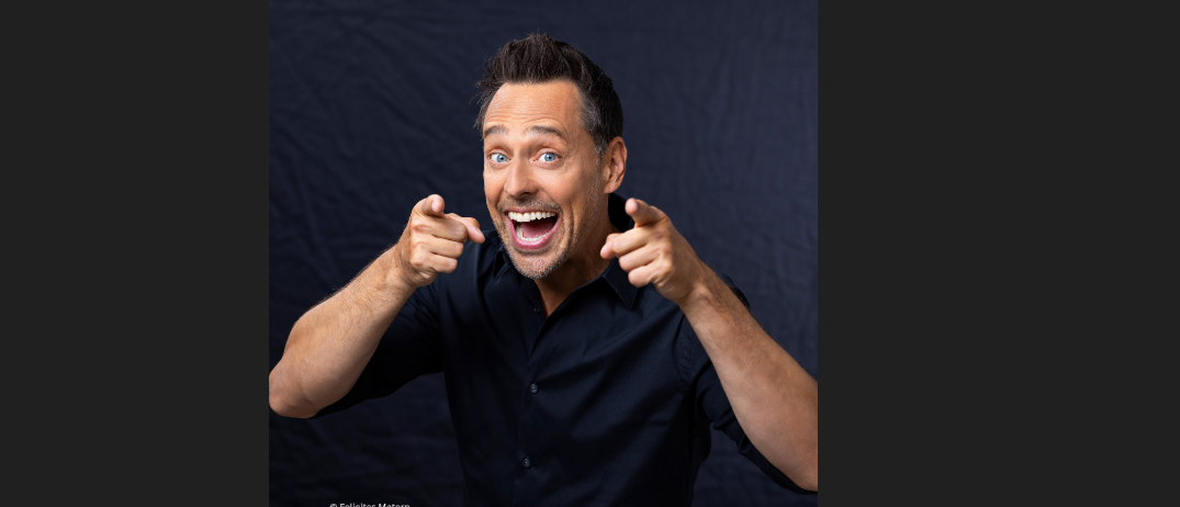 Ein Mann mit breitem Lächeln und Zeigefingern posiert für ein Foto in einem schwarzen Hemd.