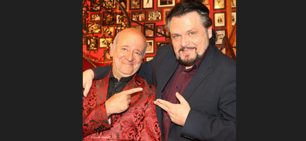 Zwei Männer stehen nebeneinander, einer trägt einen rot gemusterten Mantel und der andere ein dunkles Kostüm, und posieren für ein Foto. Sie lächeln beide und scheinen für eine Kamera zu posieren.