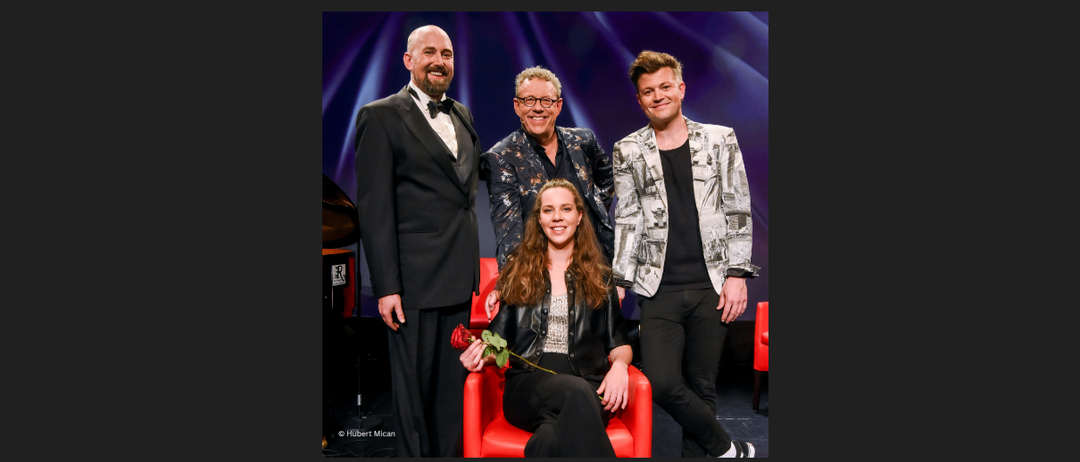 Vier Personen, drei Männer und eine Frau, posieren für ein Foto auf der Bühne mit blauem Hintergrund. Die Frau sitzt auf einem roten Stuhl und hält eine Rose. Die Männer stehen hinter ihr, einer trägt einen Fliege.