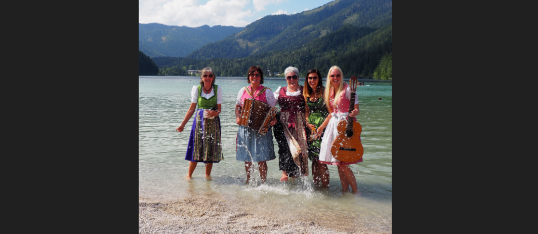 Fünf Frauen stehen im See, lächelnd, mit Bergen und einem See im Hintergrund. Jede hält ein Musikinstrument, eines eine Ziehharmonika und andere Gitarren.