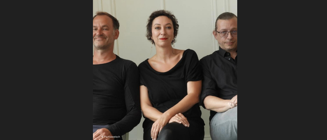 Drei Personen sitzen nebeneinander, lächeln und scheinen für ein Foto zu posieren. Alle drei sind in Schwarz gekleidet.