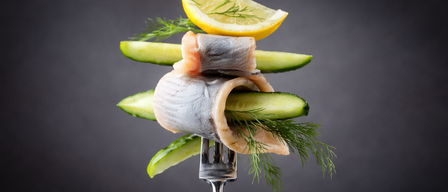 Eine Gabel mit Räucherfisch, Gurkenscheiben und einer Zitronenscheibe, garniert mit Dill auf dunklem Hintergrund.