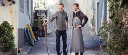 Zwei Männer in Arbeitskleidung stehen auf einer engen Straße, einer hält eine Harke und lächelt, während sie für ein Foto posieren.