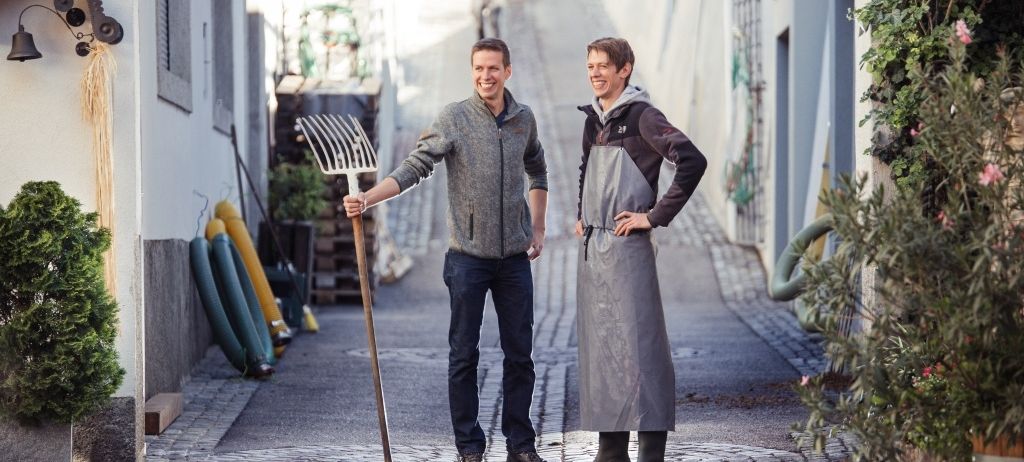 Zwei Männer in Arbeitskleidung stehen auf einer engen Straße, einer hält eine Harke und lächelt, während sie für ein Foto posieren.