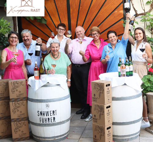 Eine Gruppe von Menschen lächelt und hält Weingläser in der Hand, sie stehen vor Holzfässern. Sie feiern wahrscheinlich ein Ereignis. Einige von ihnen tragen Brillen und einer trägt eine Uhr. Sie stehen vor einer Holzwand mit einem Logo. Es gibt auch Weinkartons und Topfpflanzen.