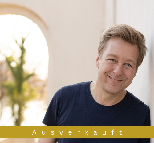 Ein lächelnder Mann in einem blauen T-Shirt lehnt sich an eine Wand, mit einem unscharfen Bogen und Baum im Hintergrund. Die Worte 'Ausverkauft' sind über ihm angezeigt.
