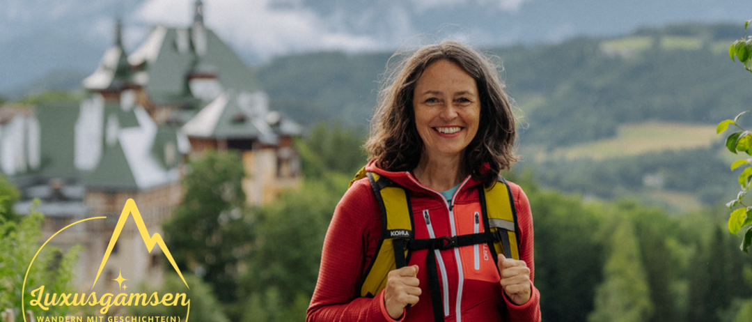 Eine Frau in einem roten Jacket und einem gelben Rucksack lächelt vor einer Berglandschaft.