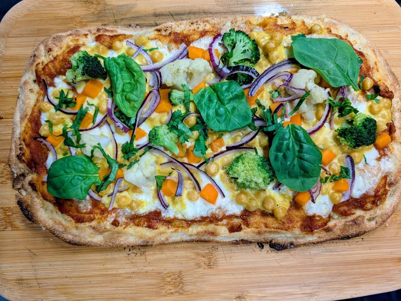 Eine große Pizza mit geschmolzenem Käse, Kichererbsen, Brokkoli, Blumenkohl und Spinat, mit roten Zwiebeln auf einem Holzschneidbrett.