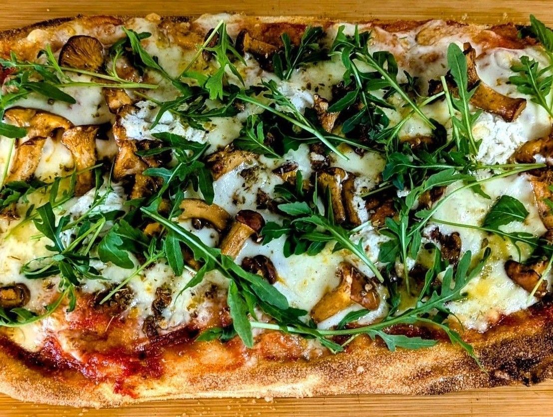 Eine Pizza mit geschnittenen Pilzen, Käse und grünen Blättern, wahrscheinlich Rucola, liegt auf einer Holzoberfläche.